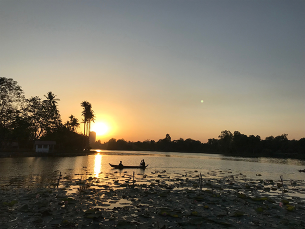 Sunset over a stretch of water with silhouette of 2 men in a boat and palm trees on the riverside